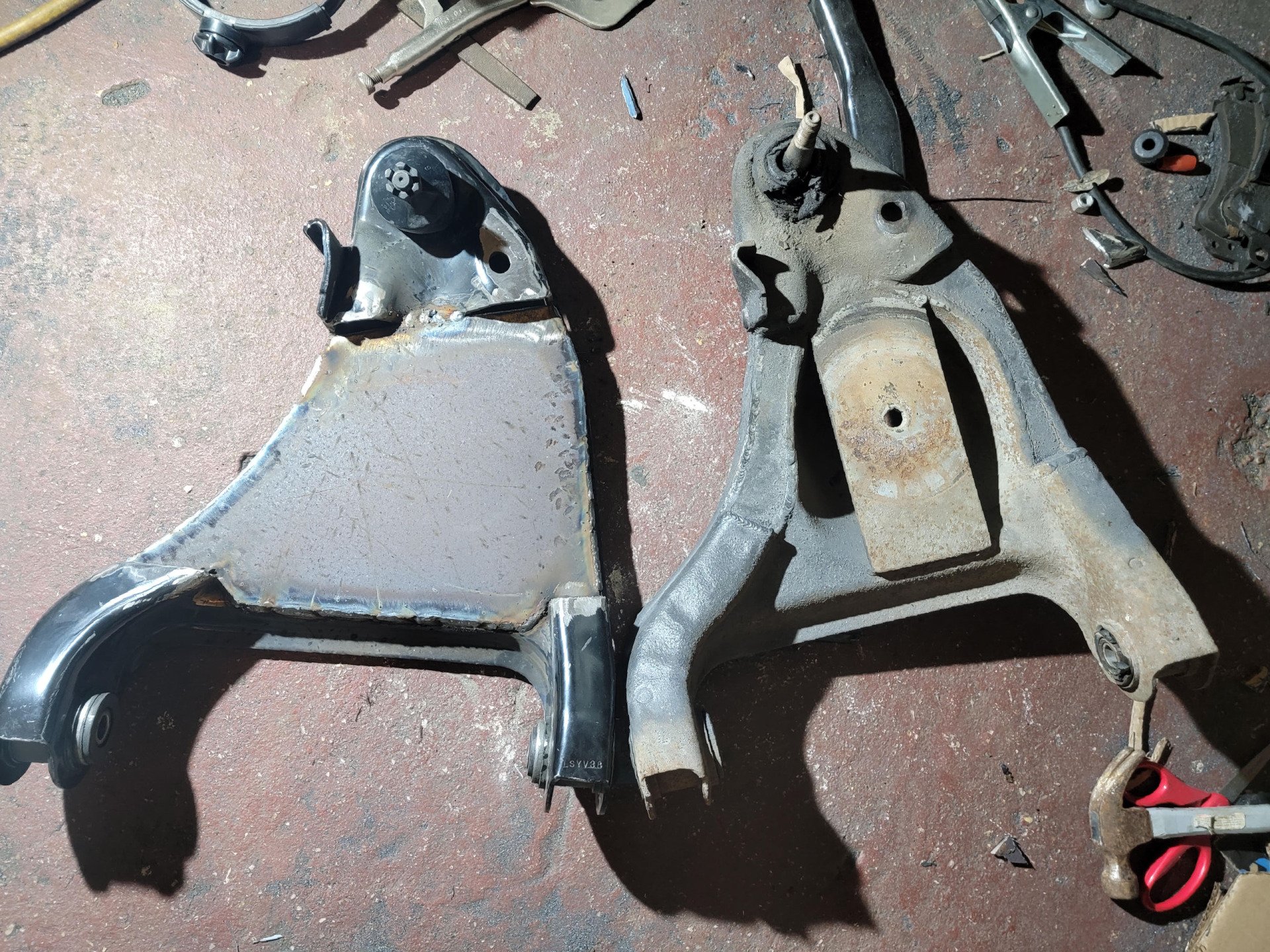 Two rusty automotive suspension control arms, one new and one old, on a workshop floor.
