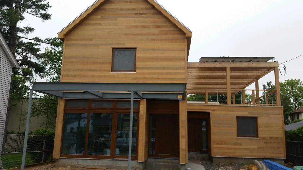 Two-story wooden house with a small patio and solar panels, viewed from the front on a cloudy day.