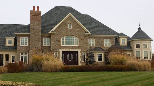 Large brick and stone house with a dark roof and decorative tower, set on a green lawn with tall grass.
