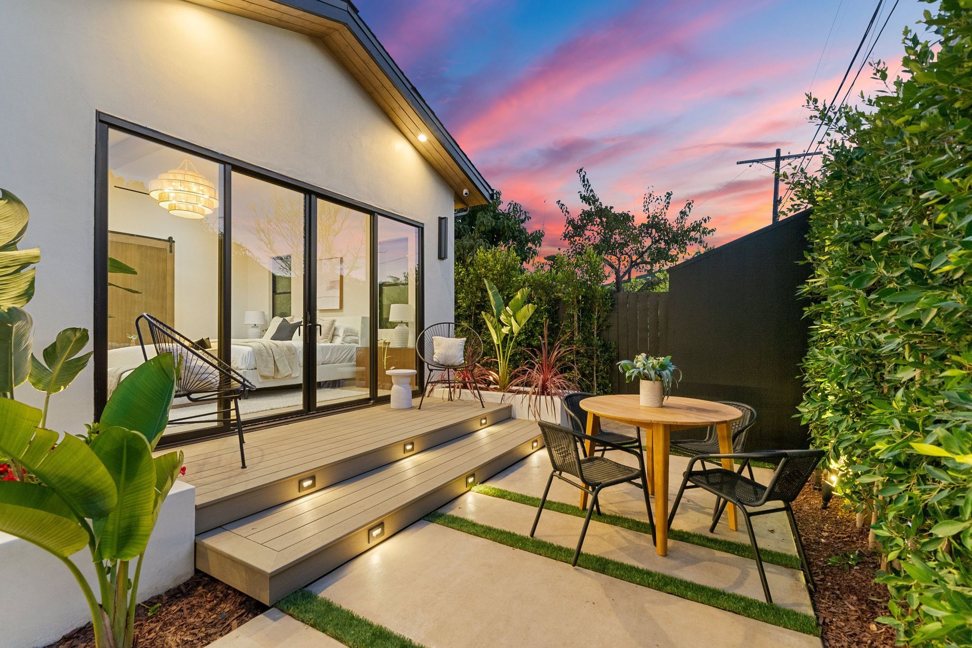 Modern patio with sliding glass doors, steps, outdoor dining table, and sunset sky beside a lit home exterior