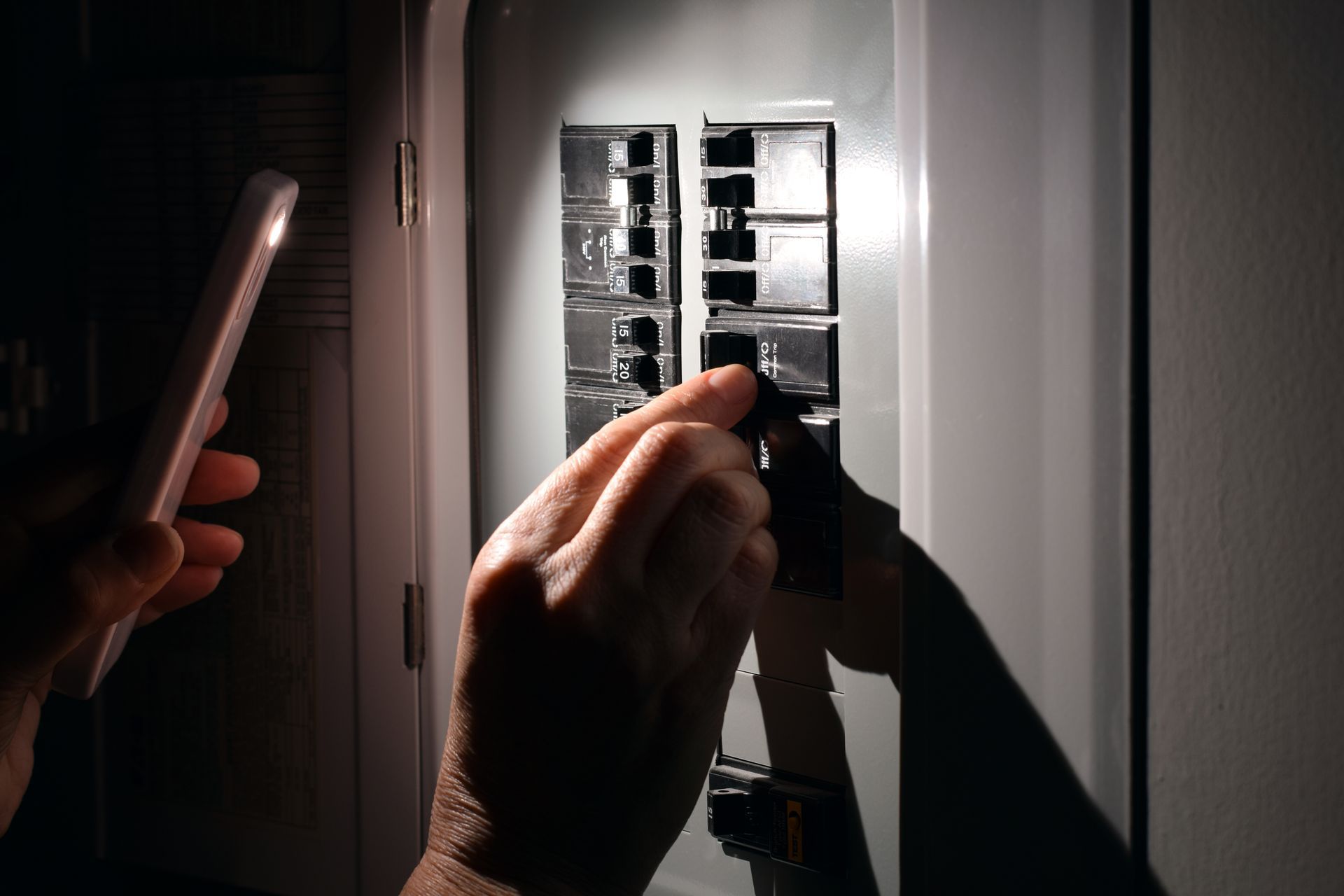 Hands working in an electrical breaker panel with a flashlight in a dim room