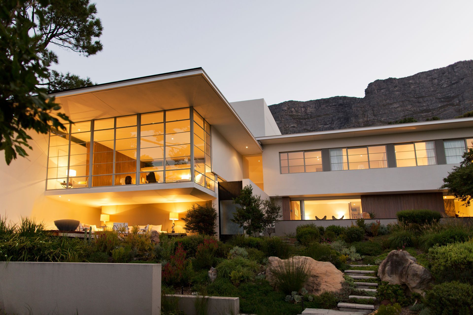 Modern house at dusk with warm interior lights, large windows, and landscaped garden in front.