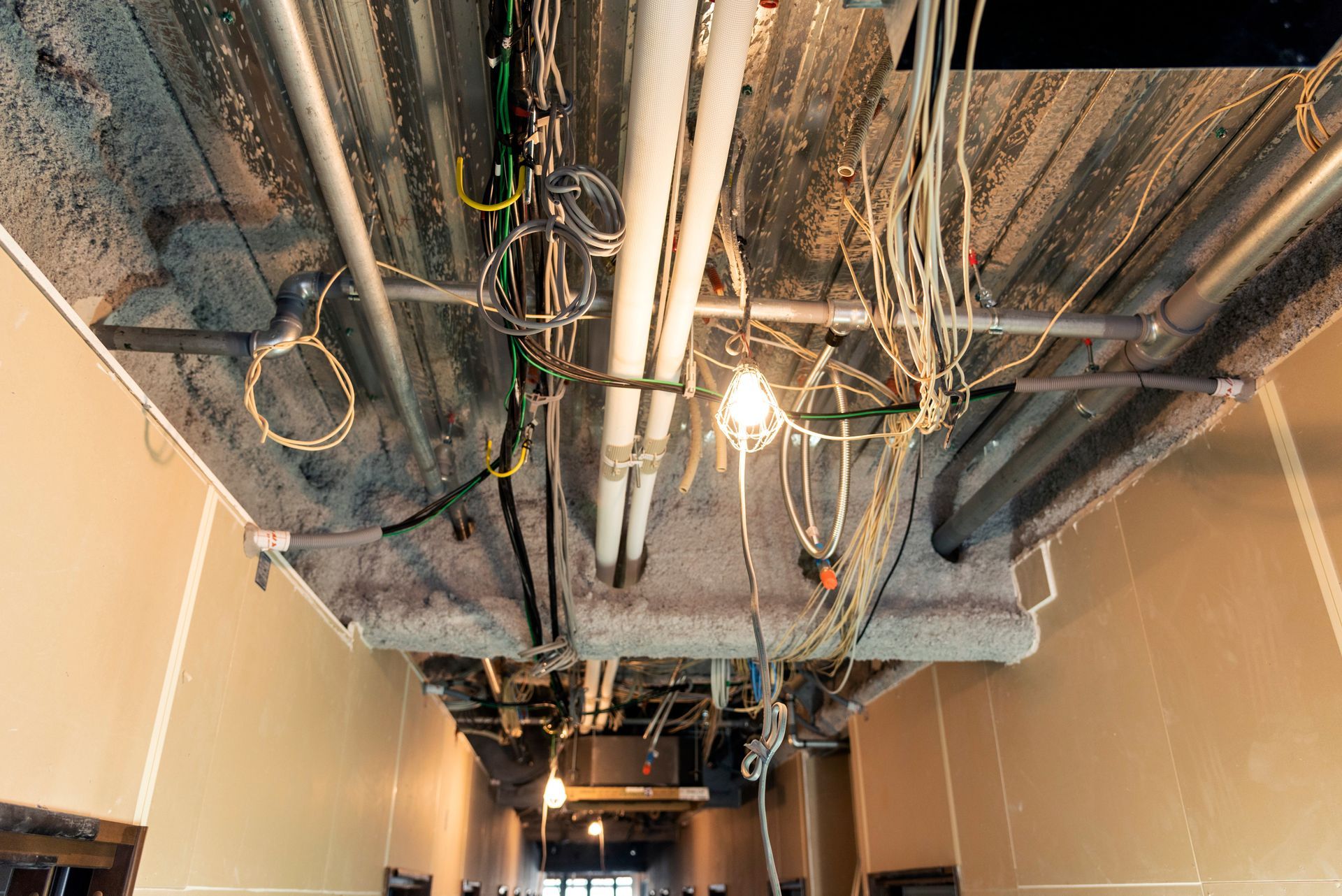 Exposed ceiling with hanging cables, pipes, and a lit bulb in an unfinished hallway