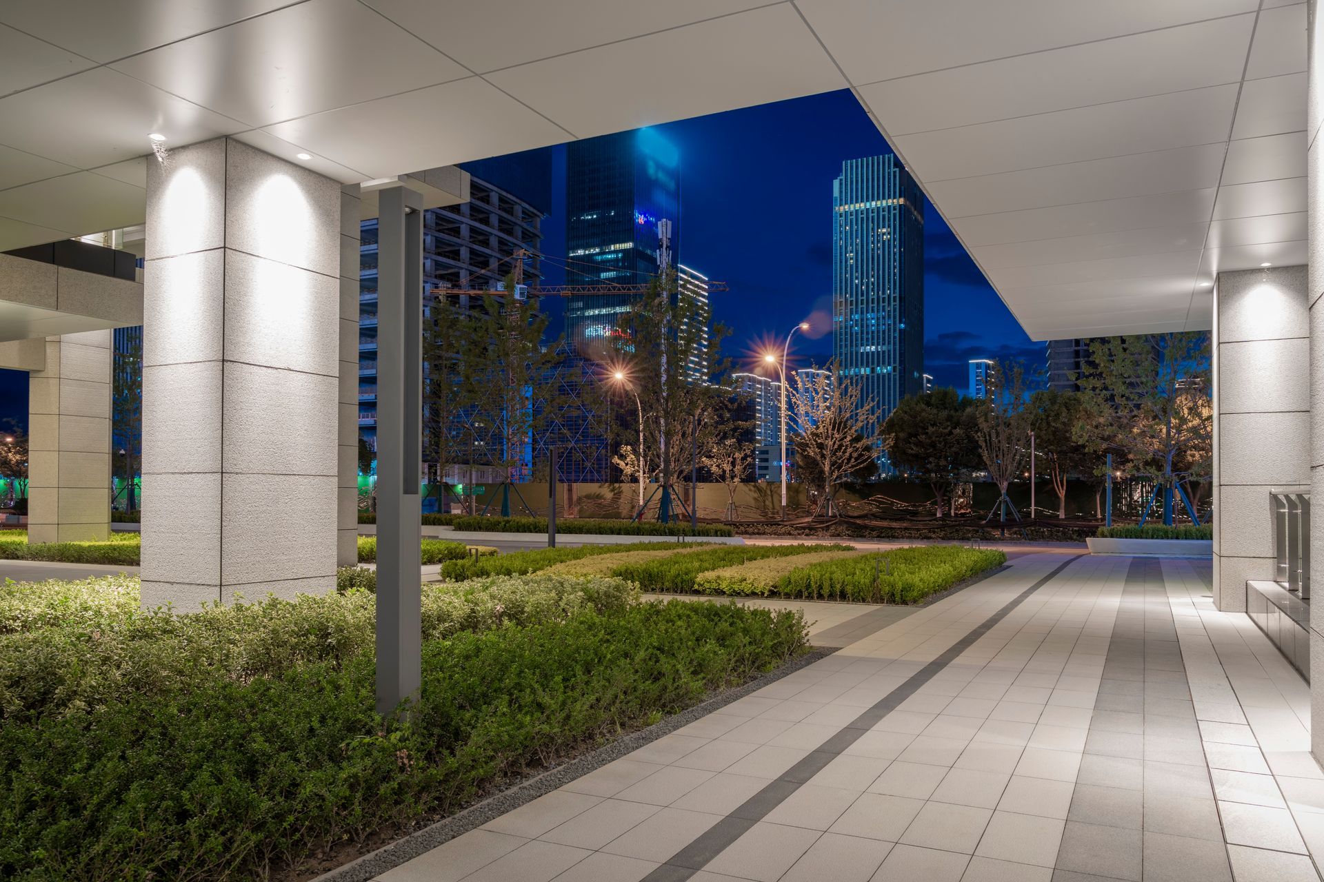 Covered plaza walkway with columns and landscaped greenery at night, city buildings glowing in the background