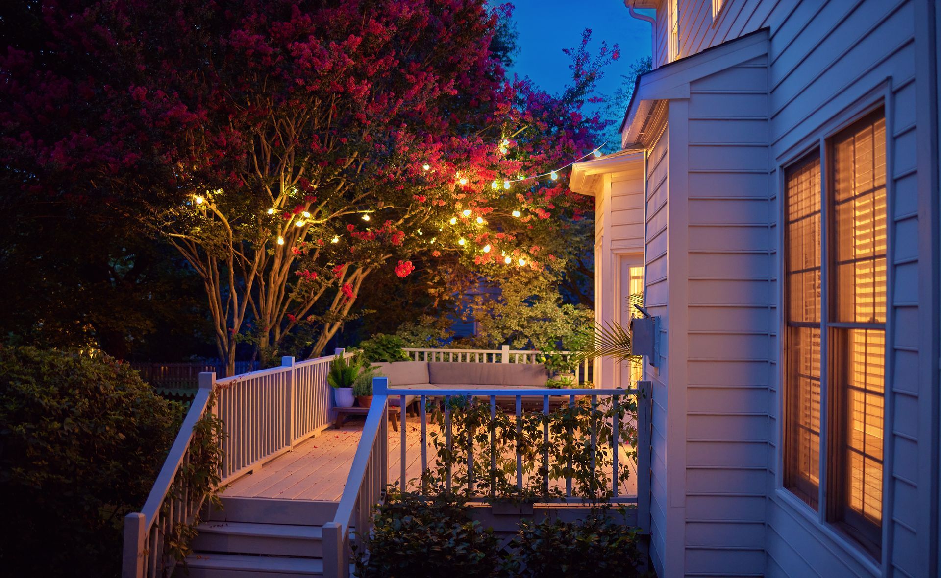 Front porch with white siding and warm lights, overlooking a lit garden at dusk