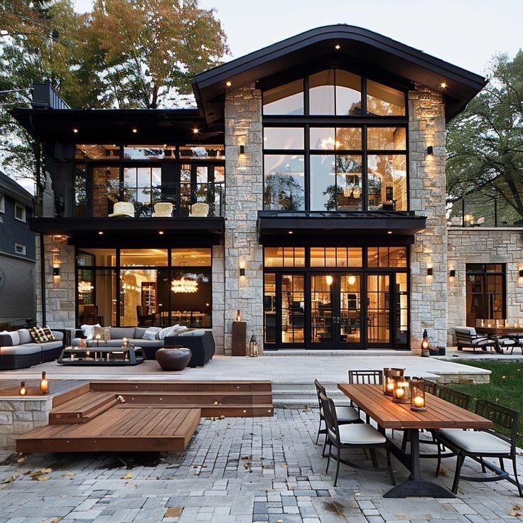 Modern two-story stone house with large windows, lit patio, and outdoor seating at dusk