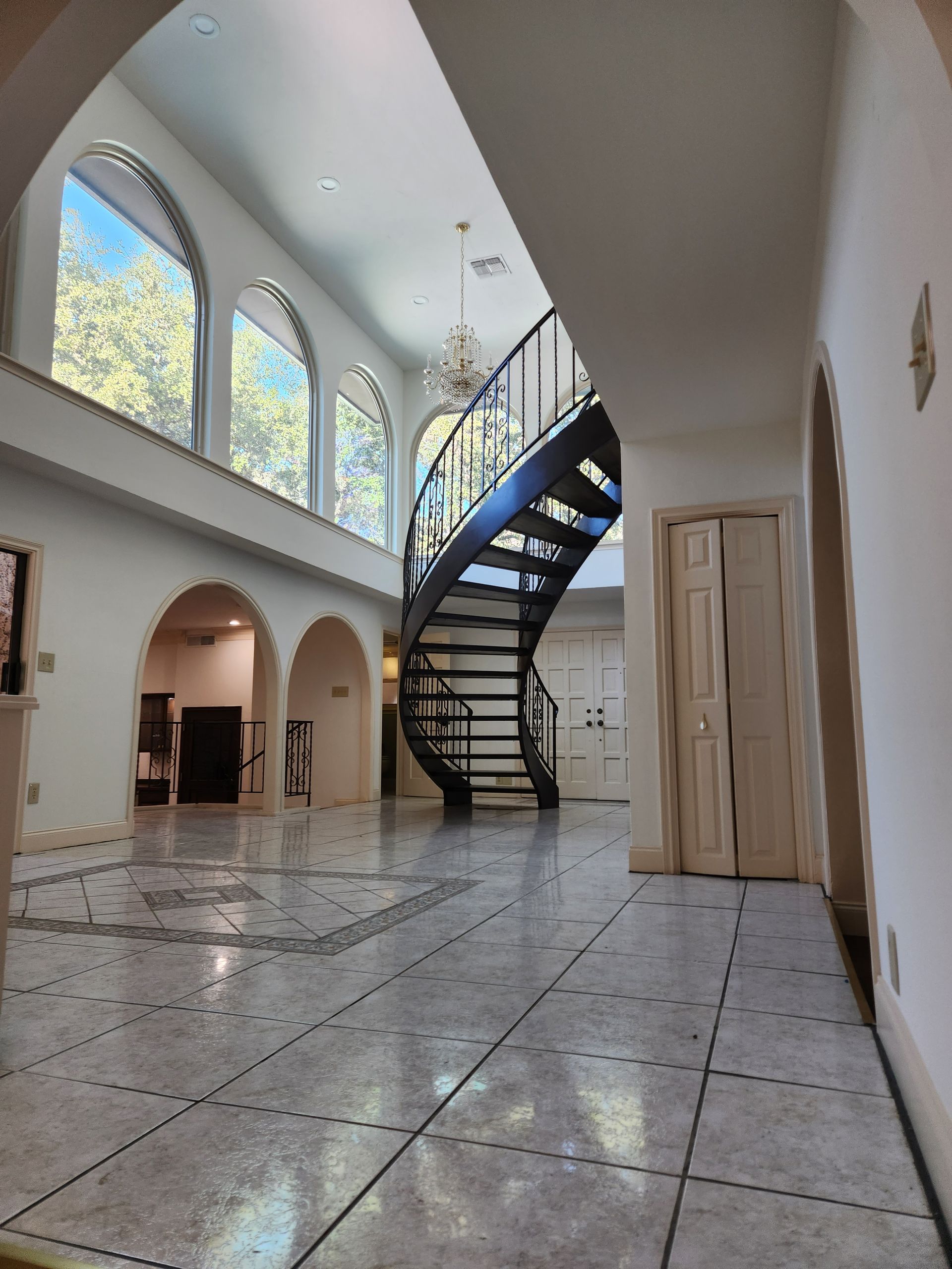 Grand foyer with tiled flooring, arched wall openings, large windows, and a curved metal staircase under a high ceiling.