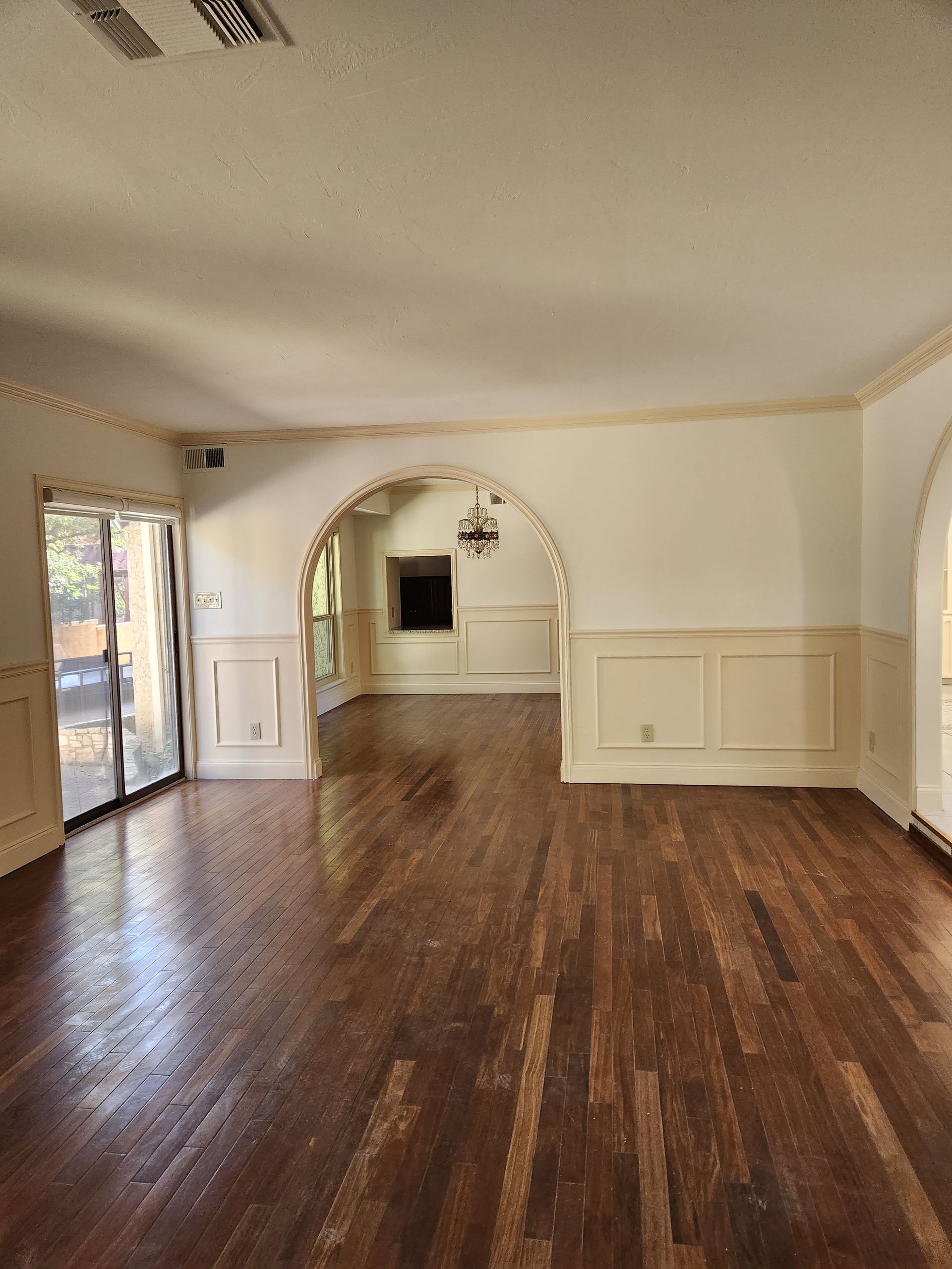 An interior view of a room with dark wood flooring, beige wainscoting, and a prominent arched entryway to another room.