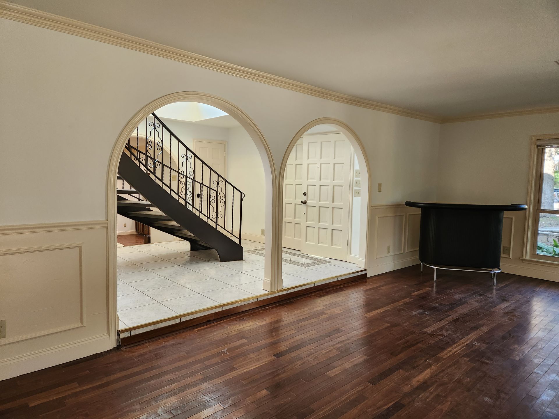 A bright room with wood floors and wainscoting features two large archways leading to a foyer with a staircase.