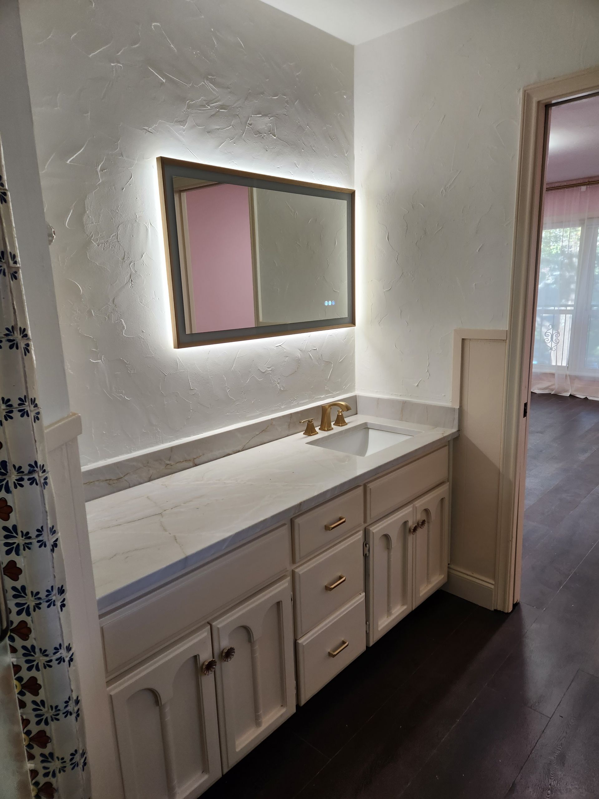 A bathroom vanity with white cabinets, a light-colored countertop, and an illuminated mirror on a textured white wall.