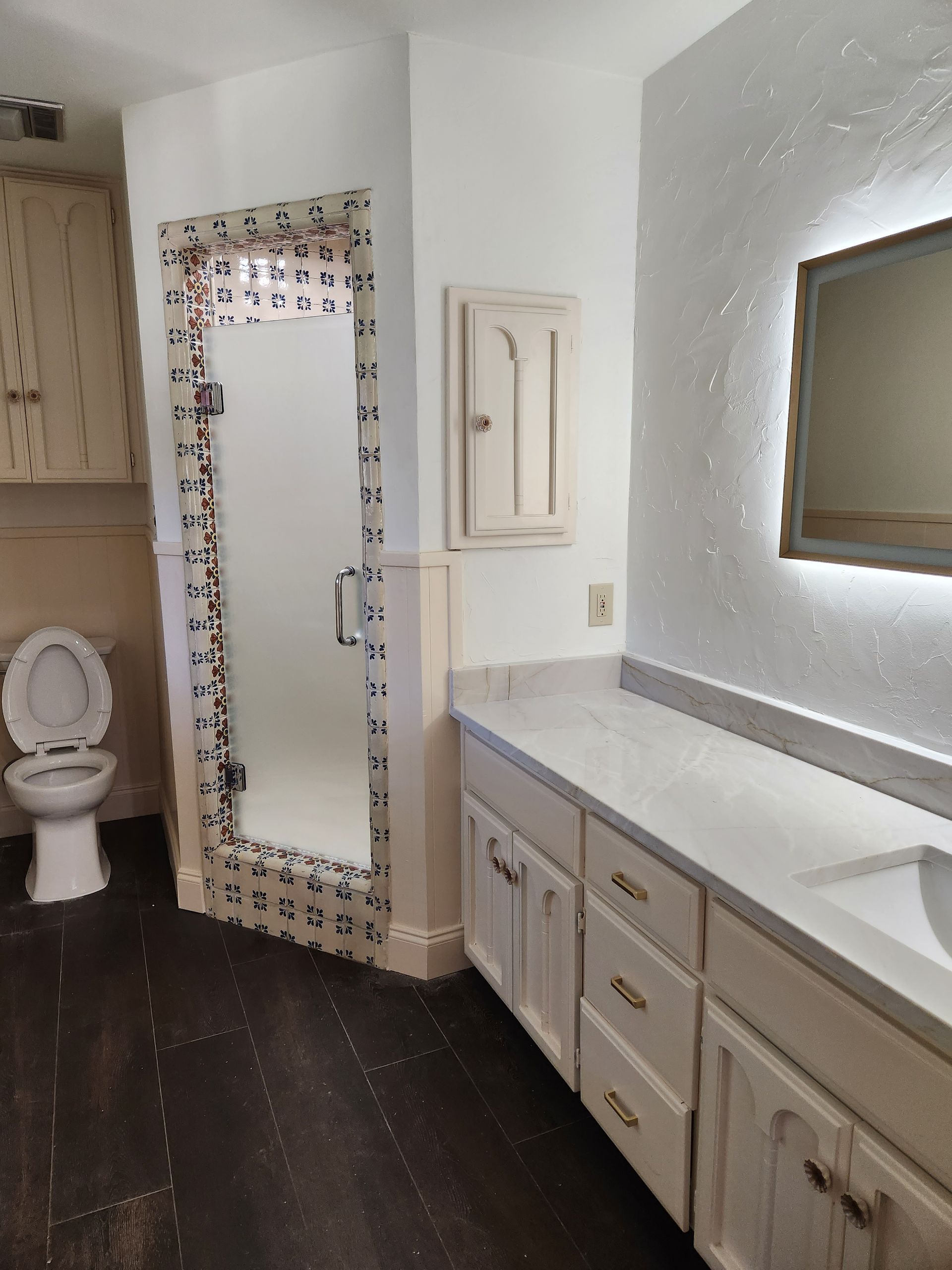 A bathroom with dark flooring, a white vanity with a lighted mirror, and a walk-in shower framed with patterned tiles.