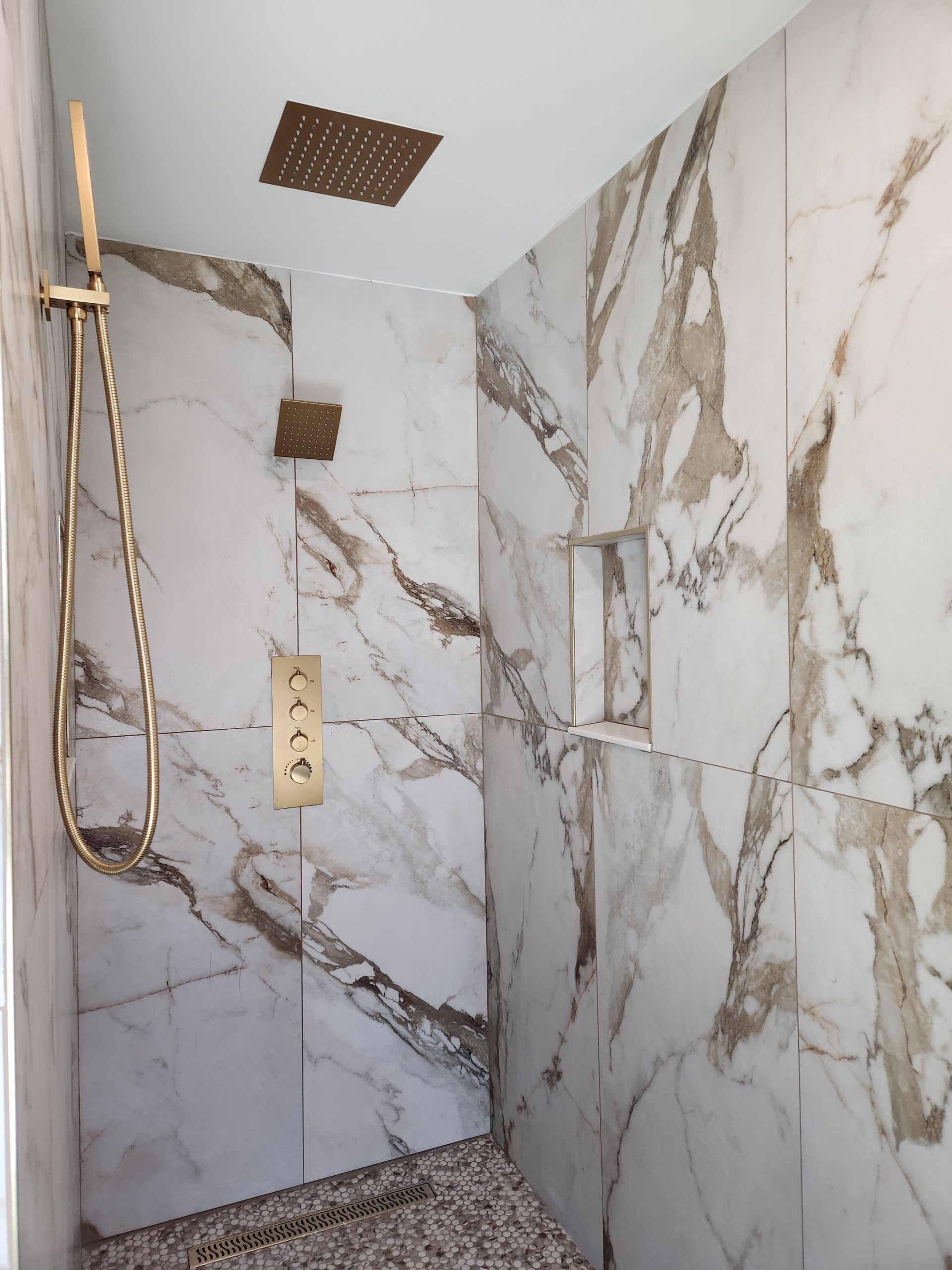 A modern walk-in shower with gold fixtures, white marble-style wall tiles, a ceiling rain showerhead, and a pebble floor.