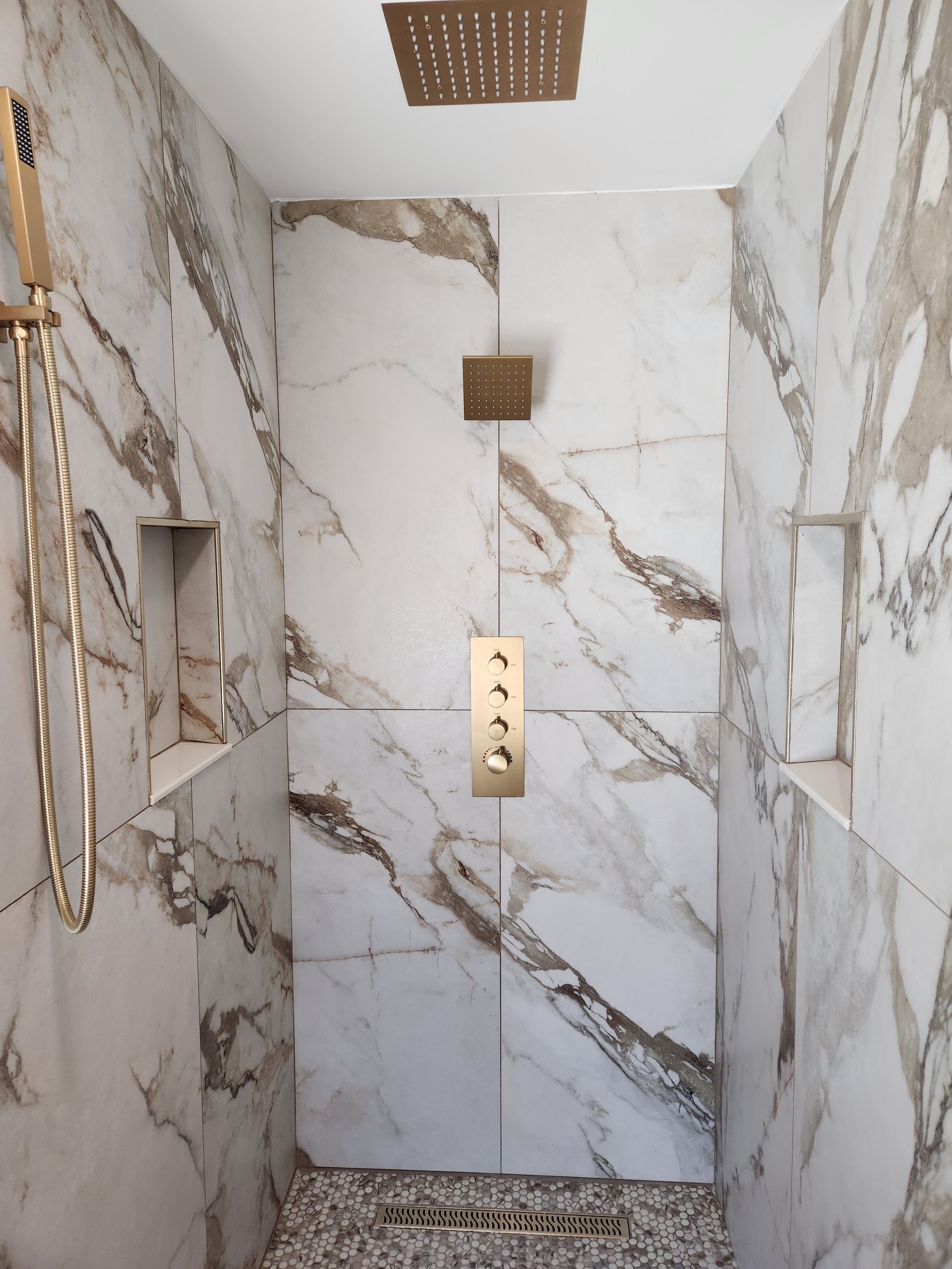 A walk-in shower with white and brown marble tiles, gold fixtures, a rain showerhead, and a pebbled floor drain.