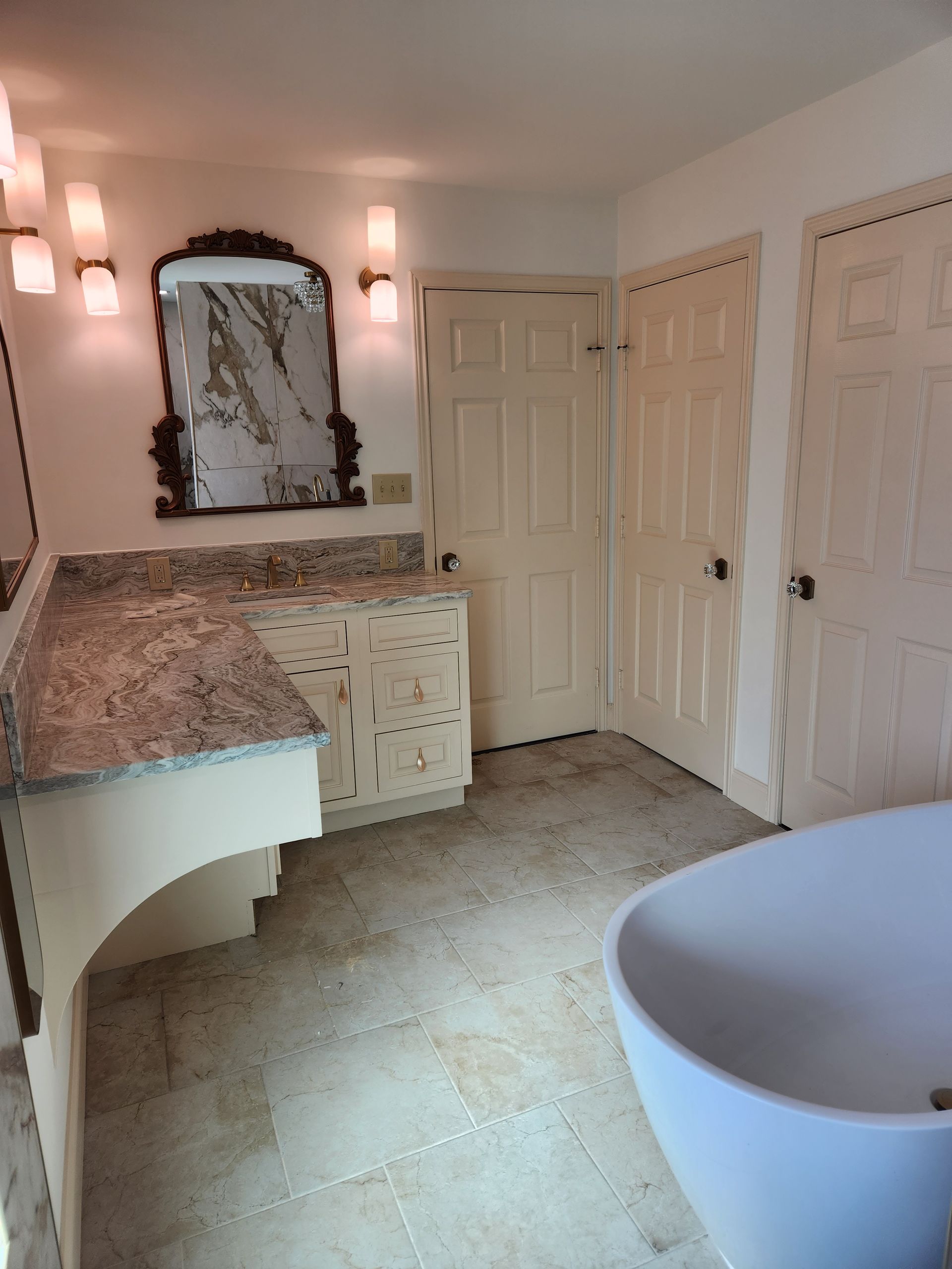 A bathroom with a granite-topped vanity, decorative mirror, two light sconces, tile flooring, and a white soaking tub.