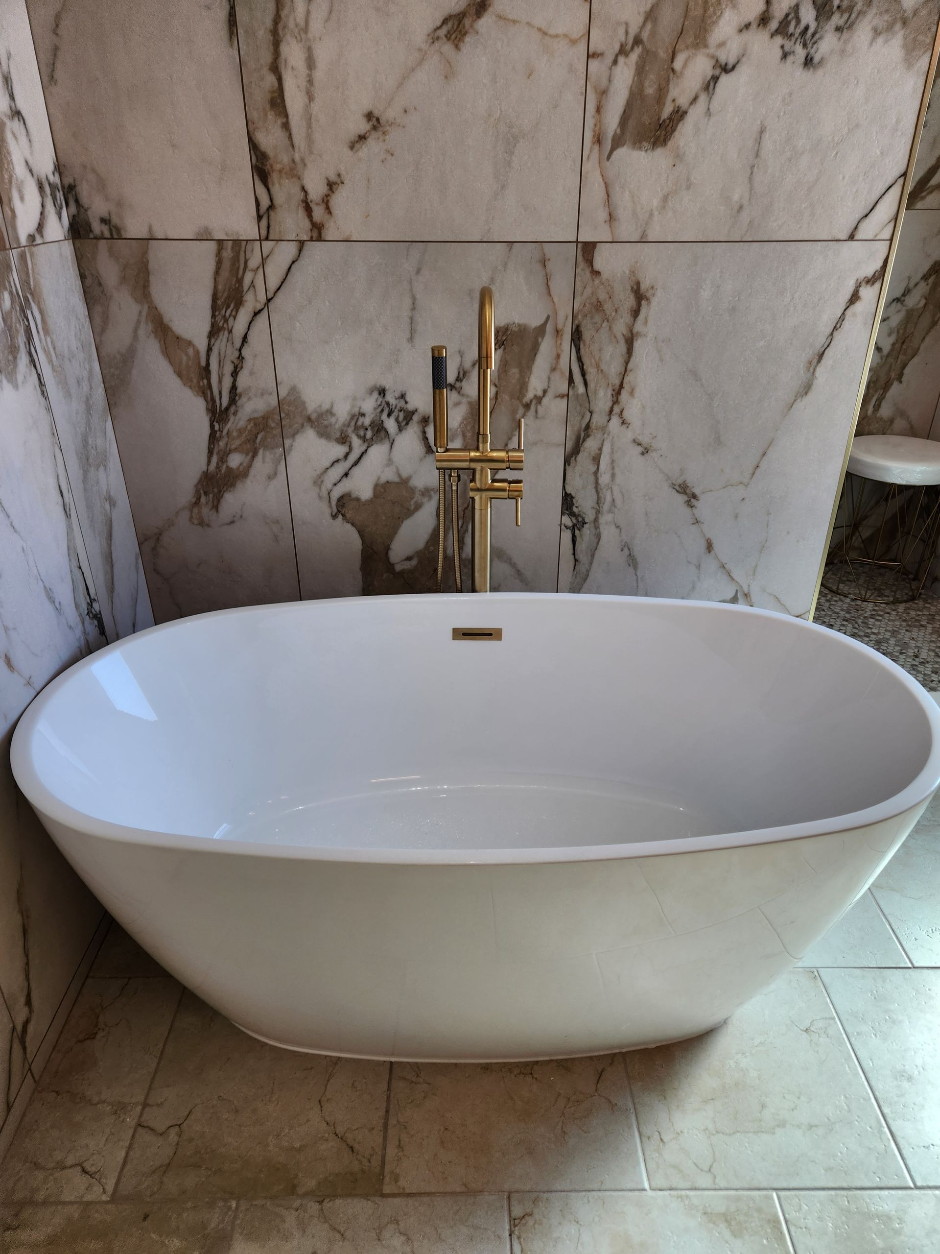A white freestanding oval bathtub with a gold floor-mounted faucet in front of a marble-tiled wall.