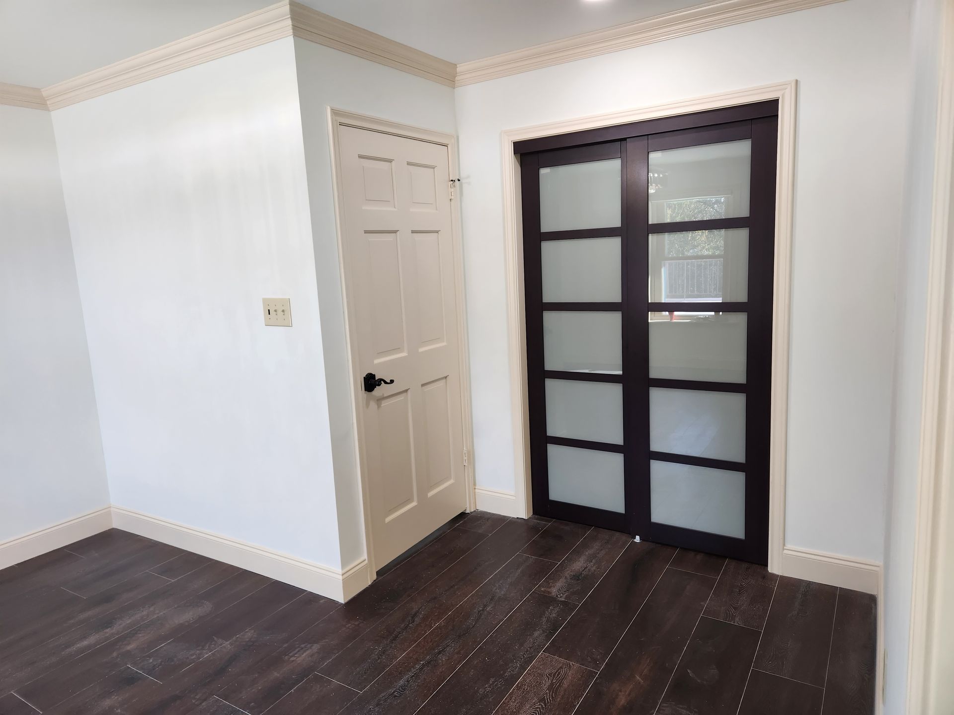 A room with dark wood flooring, white walls, crown molding, a cream-colored door, and a dark-framed sliding glass door.