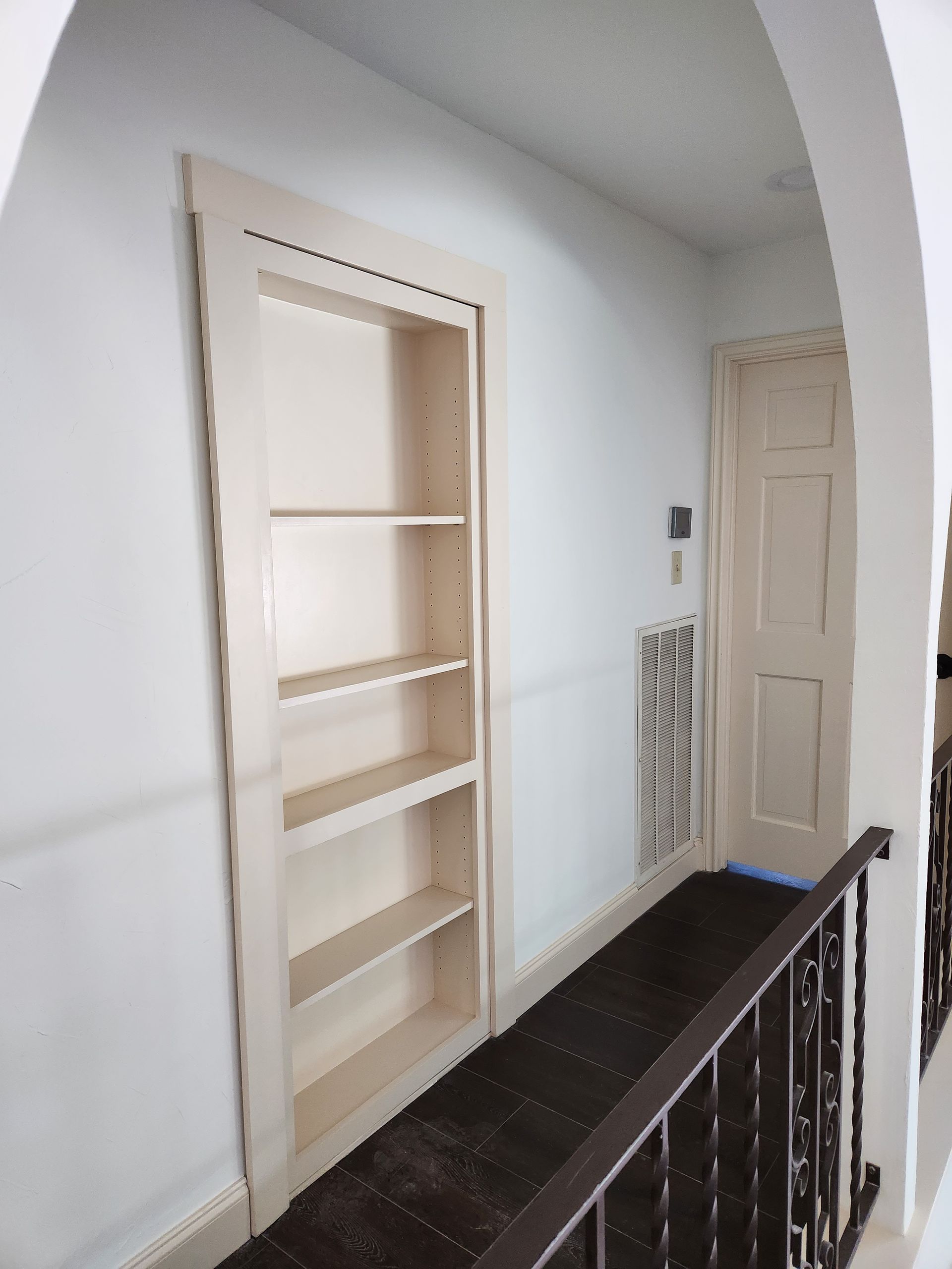 A hallway with white walls featuring an inset bookshelf, an ornate wall vent, a closed door, and a decorative metal railing.