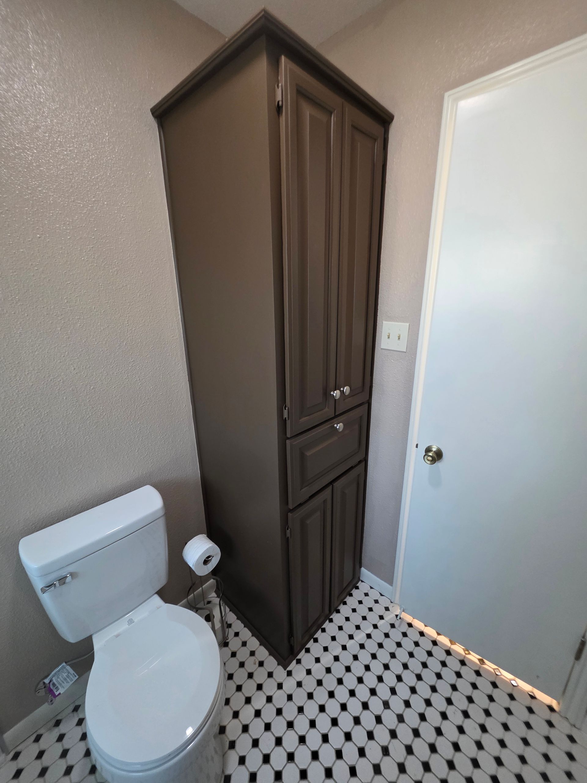 A tall, dark-brown storage cabinet next to a white toilet in a bathroom with patterned black and white floor tiles.