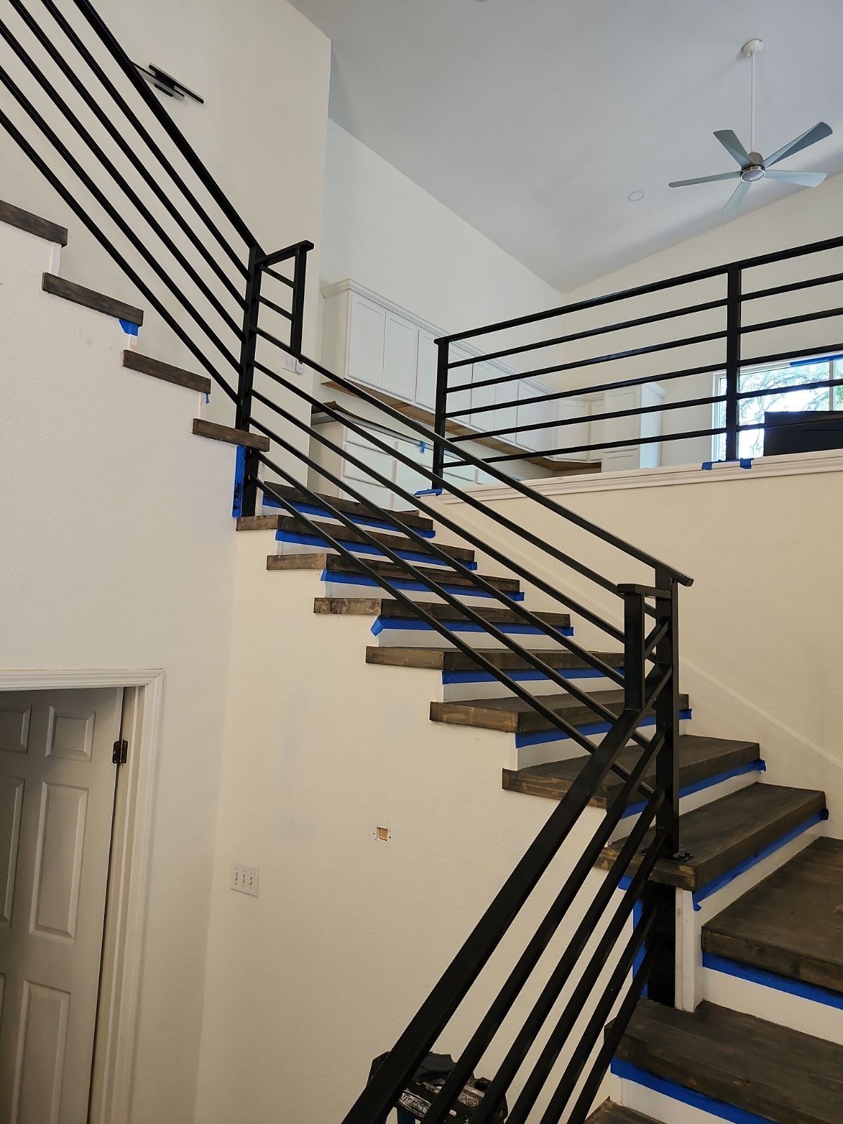 A staircase featuring dark wood treads, white risers with blue painter's tape, and a modern black metal horizontal railing.