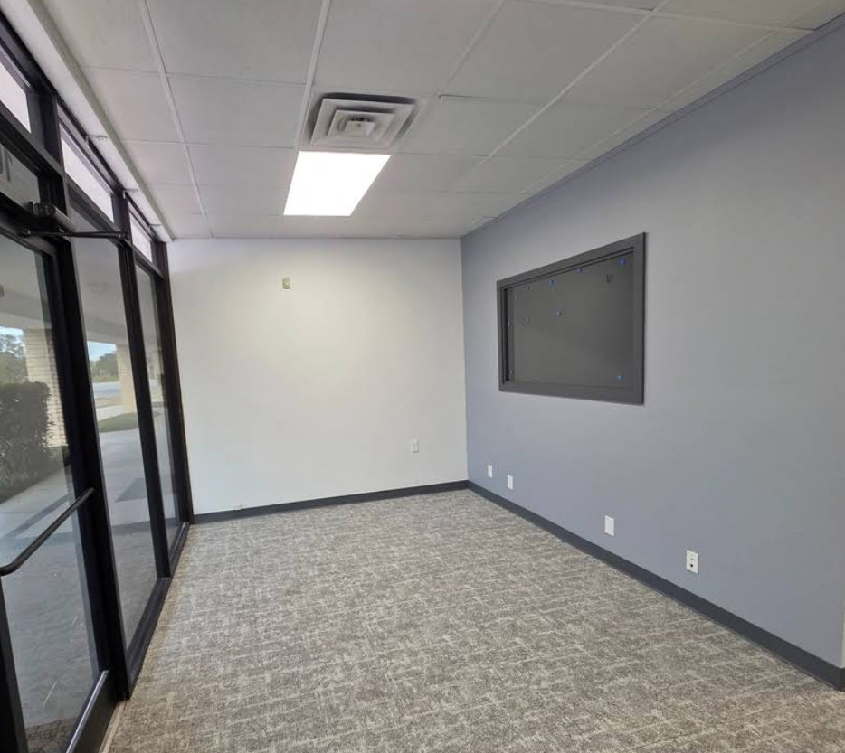 Empty office space with grey and white walls, carpeted flooring, glass entry doors, and a ceiling light fixture.