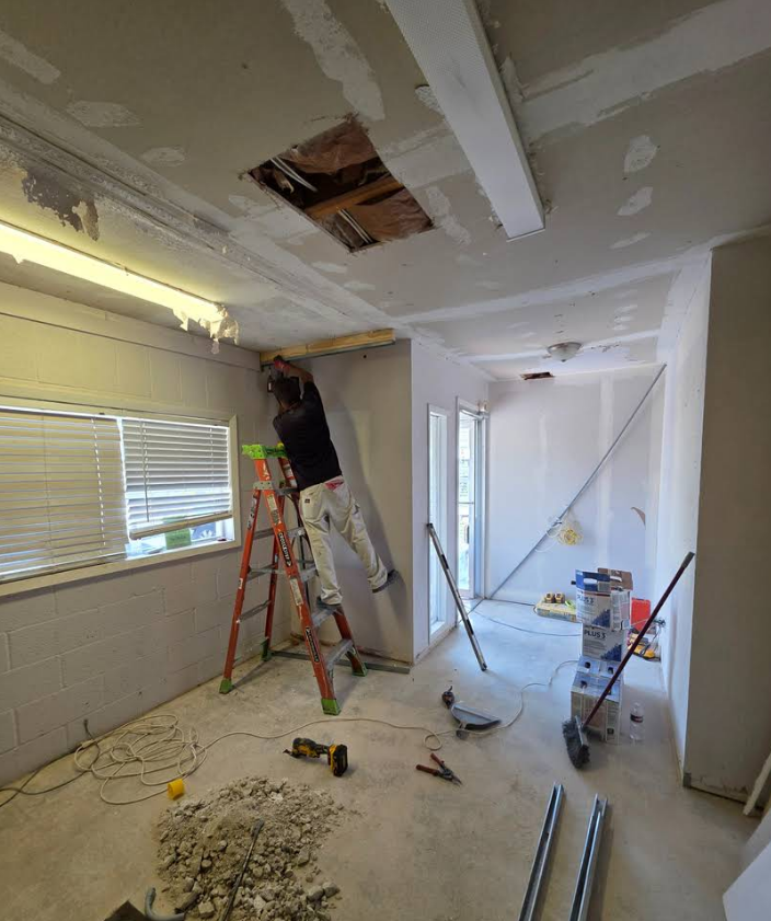 A person on a stepladder works on the ceiling and upper wall of a room undergoing construction, surrounded by tools.