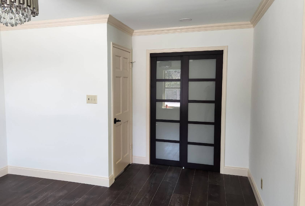 A room with white walls, a beige door, and black-framed glass double doors, all featuring crown molding and dark floors.