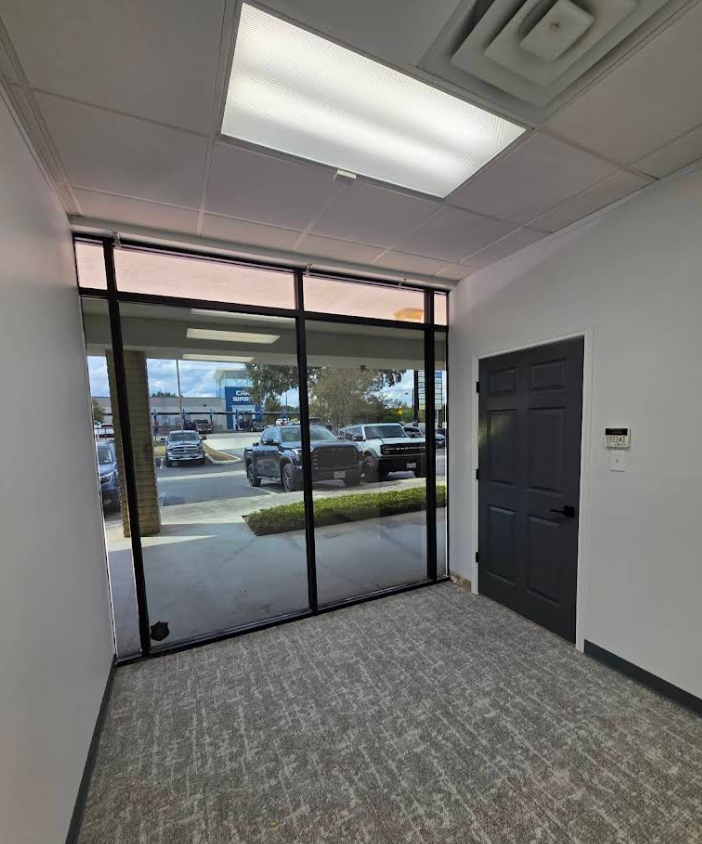 An office entryway with gray carpet, white walls, a dark door, and large windows looking out onto a parking lot.