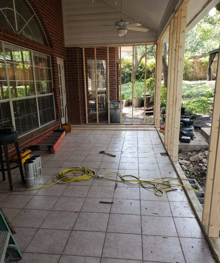 Construction of a wood-framed wall on a tiled patio, with open studs and a large window against a brick exterior wall.