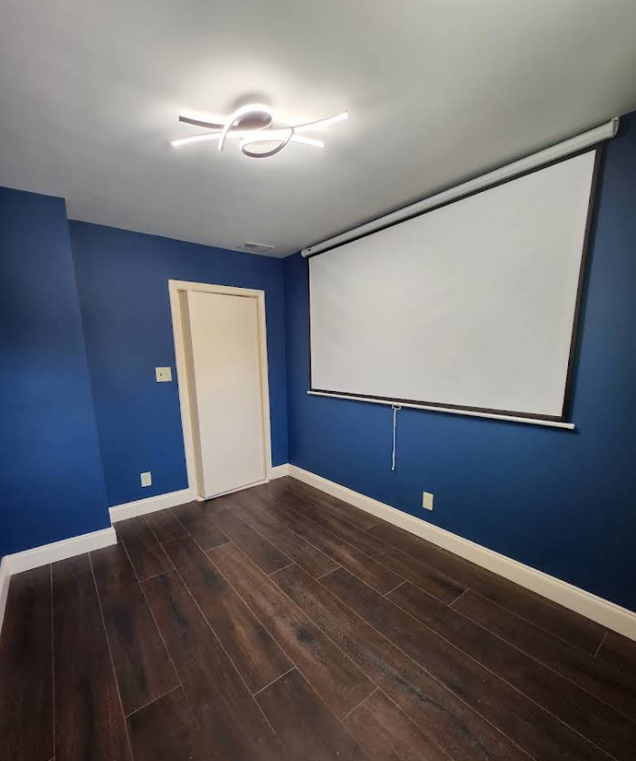 An empty room with blue walls, dark wood-look tile floors, a white projection screen, and a modern ceiling light.