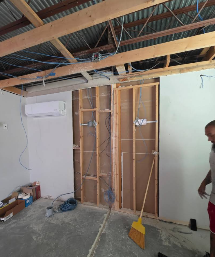 Interior room under construction with exposed wooden wall studs, ceiling beams, electrical wiring, and a mounted AC unit.