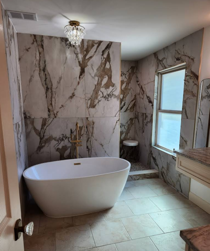 A bathroom featuring a freestanding white tub against a large-format brown and white marble accent wall and a window.