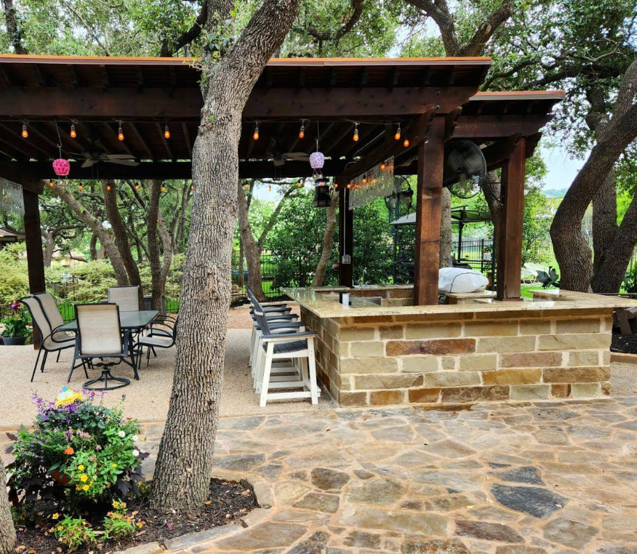 An outdoor stone patio with a wooden pergola, bar seating, a dining area, and lush green trees in a backyard setting.