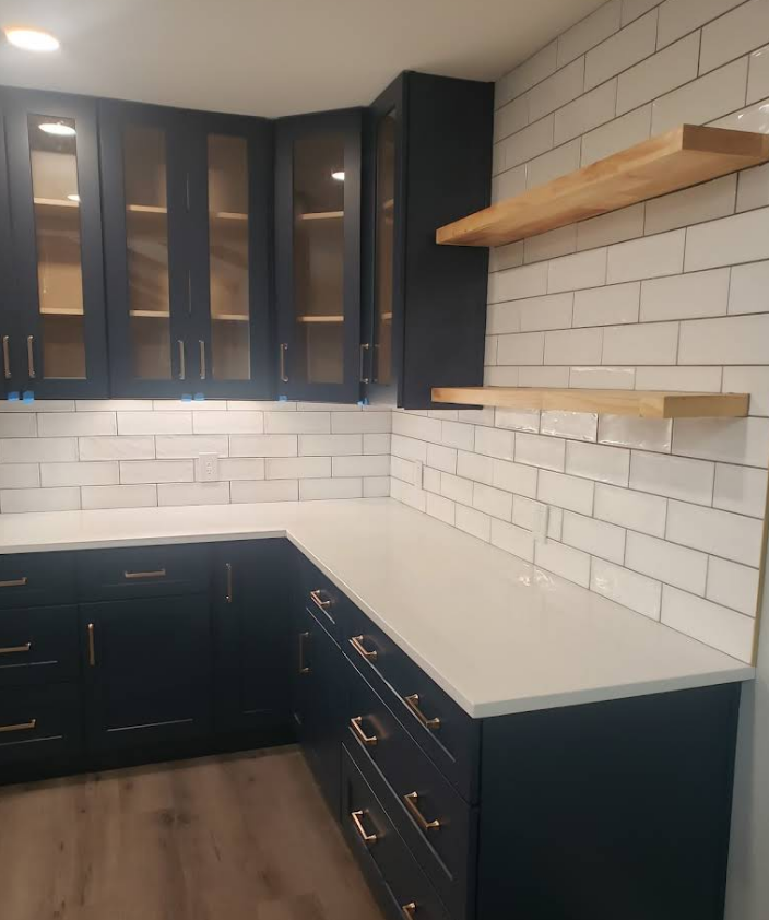 A kitchen corner with dark blue cabinets, white quartz countertops, white subway tile backsplash, and two wooden shelves.