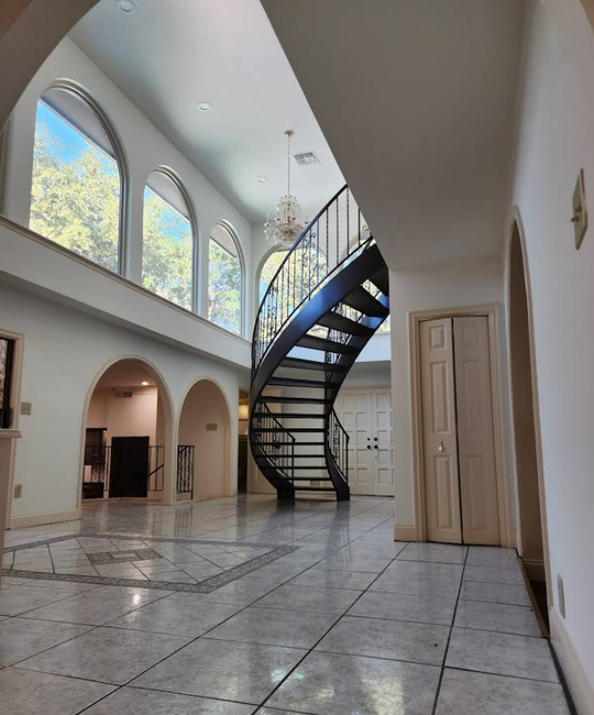 A wide foyer with light tile floors features an arched spiral staircase and three large arched windows overlooking trees.