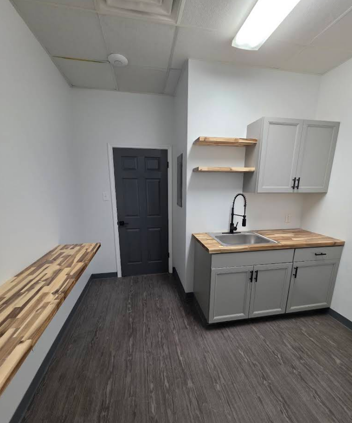 A kitchenette with gray cabinets, wood-grain counters, floating shelves, a black door, and dark flooring in a white room.