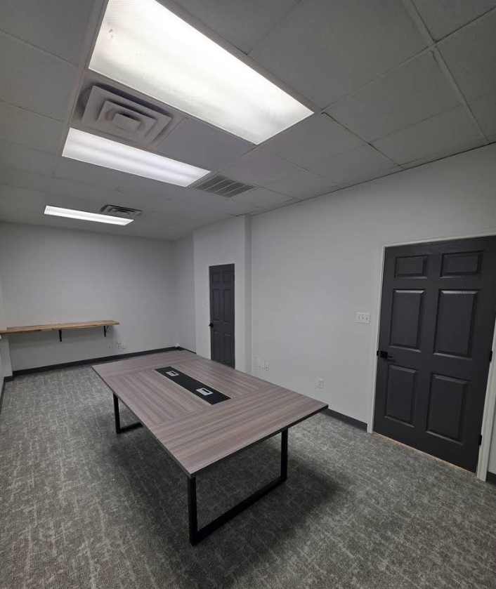 A brightly lit meeting room with a wooden table in the center, gray carpet, white walls, and two dark gray doors.