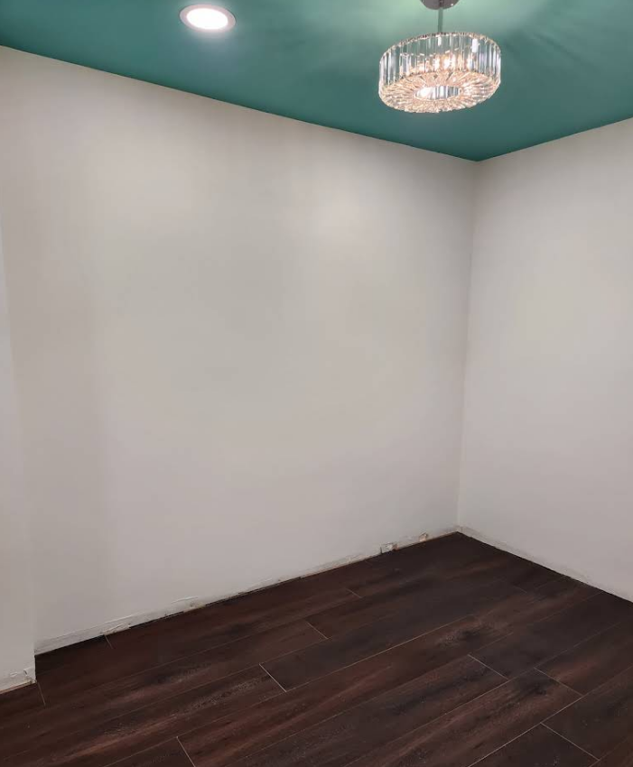 An empty corner room with plain white walls, a dark wood-look floor, and a blue ceiling with a modern crystal light fixture.