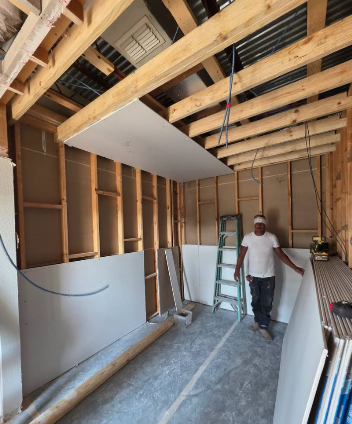 A construction worker stands in a room with exposed wooden framing, partially installed drywall, and a metal ladder.