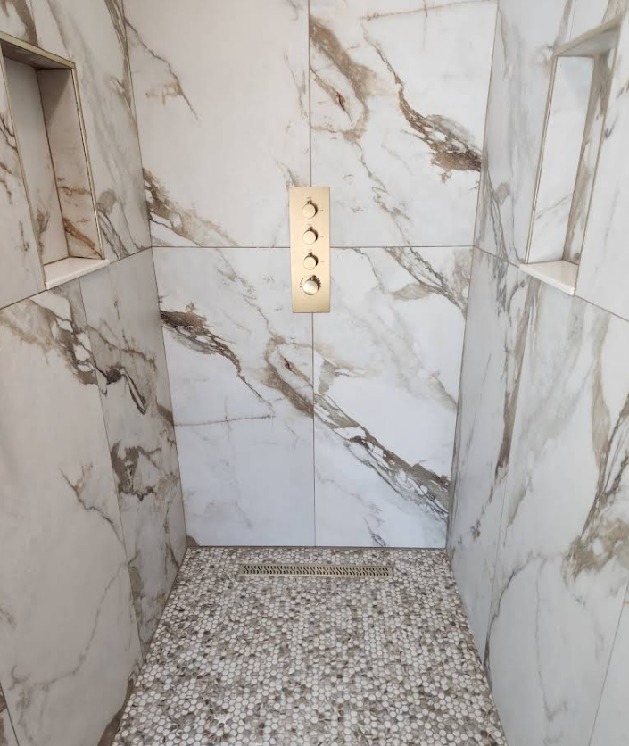 A walk-in shower with white and grey marble-patterned tiled walls, recessed shelves, a gold control panel, and pebbled floor.