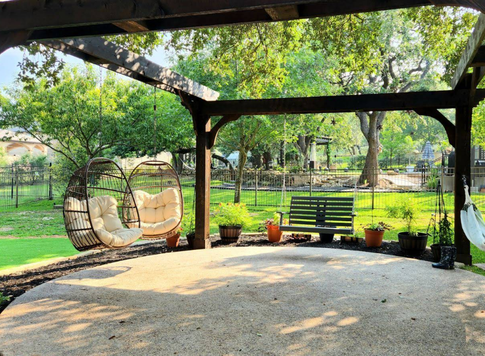 A shaded patio with two wicker egg chairs, a wooden bench, and potted plants in a sunny backyard.