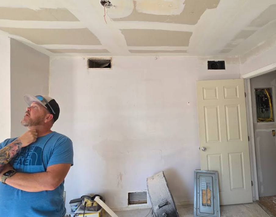 A person with a beard and baseball cap stands in a room under construction, looking up at an unfinished drywall ceiling.