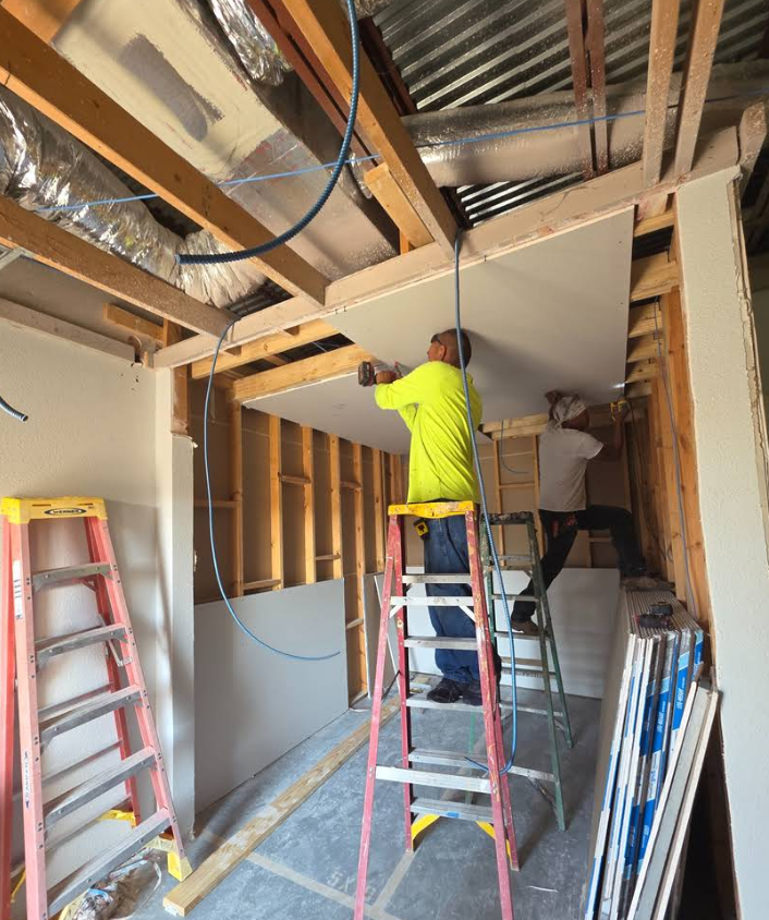 Two construction workers install a drywall ceiling panel inside an unfinished, wood-framed room with exposed ductwork.