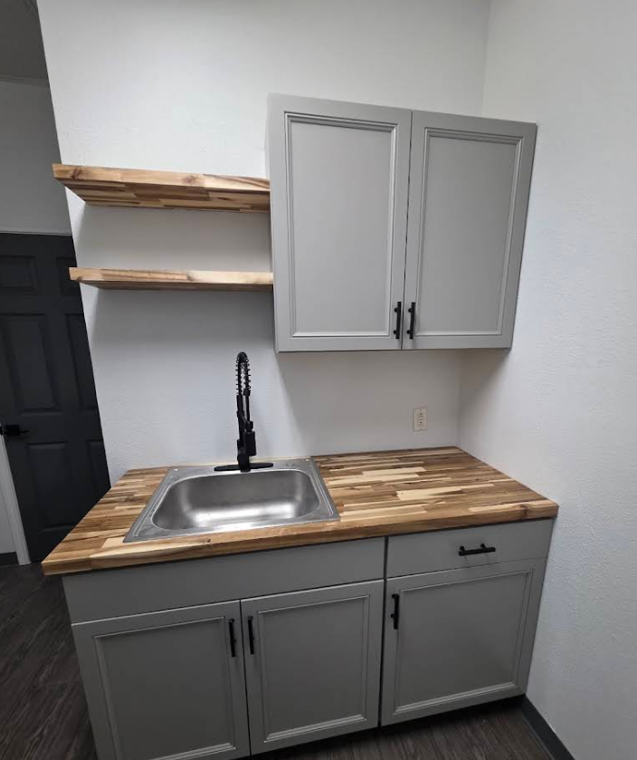 A sink area with light gray cabinets, a butcher block countertop, two matching floating shelves, and a black faucet.