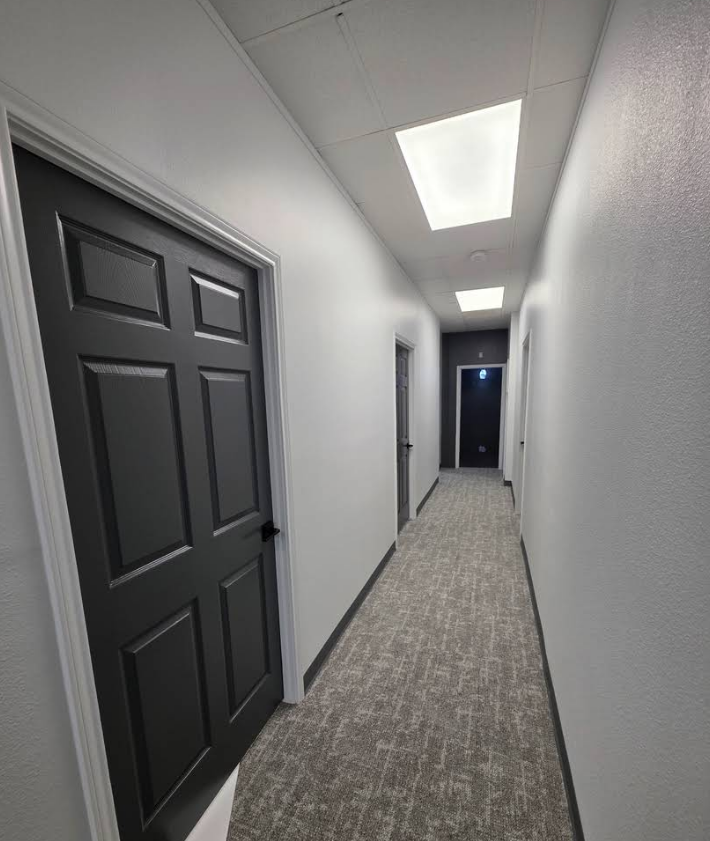 A brightly lit office hallway with light gray walls, patterned carpet, and multiple dark doors.