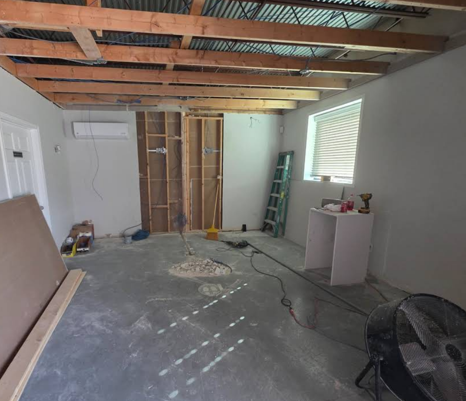 A room under construction with exposed wooden ceiling beams, unfinished drywall, a concrete floor, and a ladder.