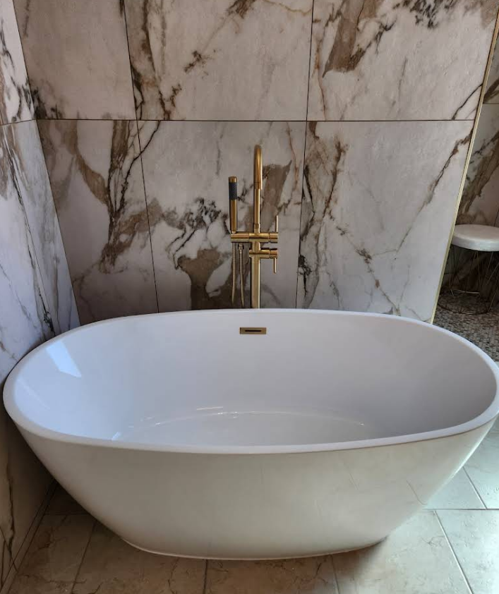 A white, oval freestanding bathtub with a gold floor-mounted faucet against a marble-tiled wall.