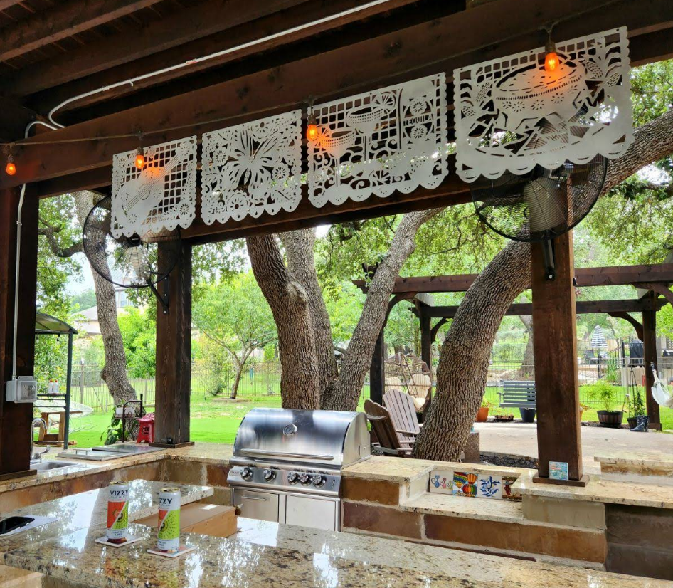 Outdoor kitchen with a built-in grill, granite counter, and white papel picado banners hanging under a wooden pergola.