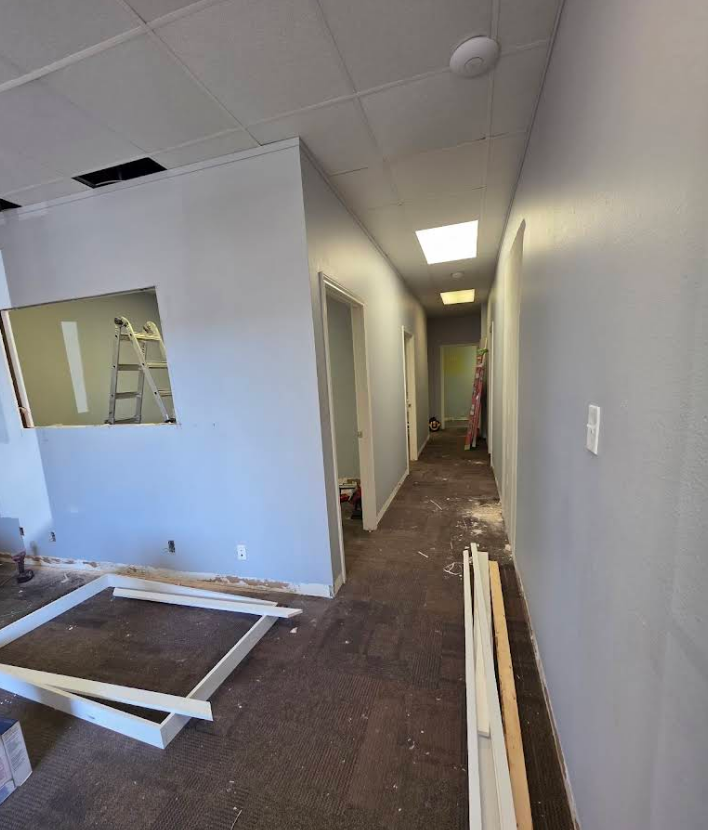 An interior view of a commercial hallway under renovation with light gray walls, dark flooring, and building materials.
