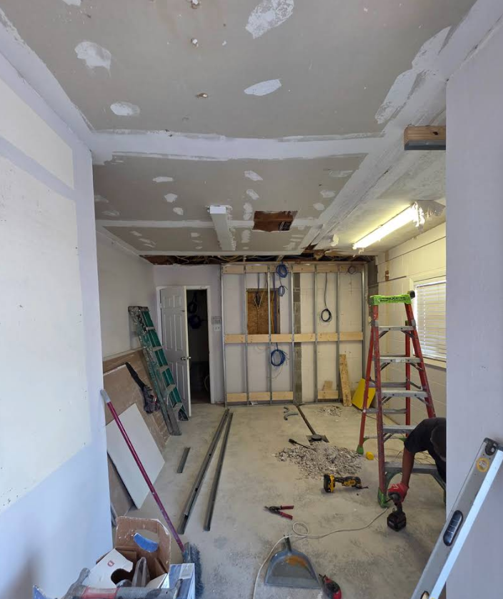 An interior room under renovation with exposed metal wall studs, plaster ceiling patches, ladders, and scattered tools.