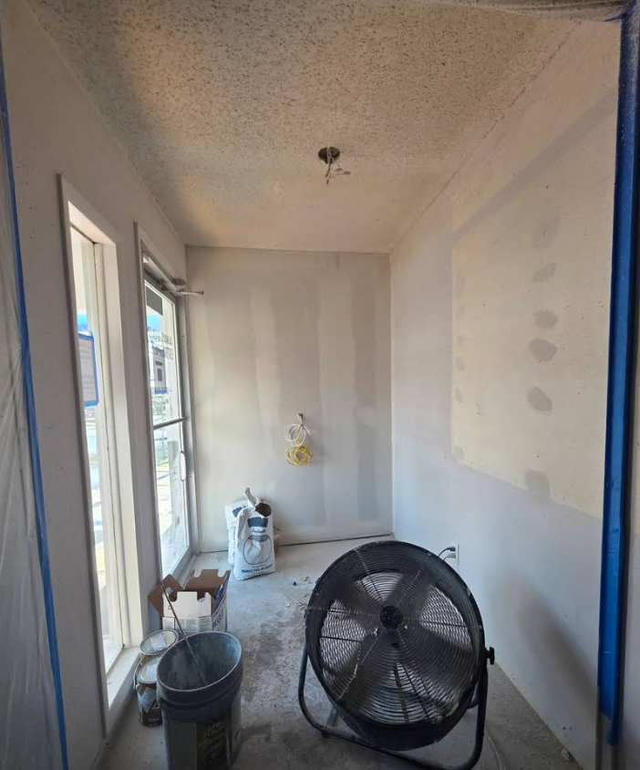 A small room under renovation with bare drywall, a popcorn ceiling, a large floor fan, and painting supplies.